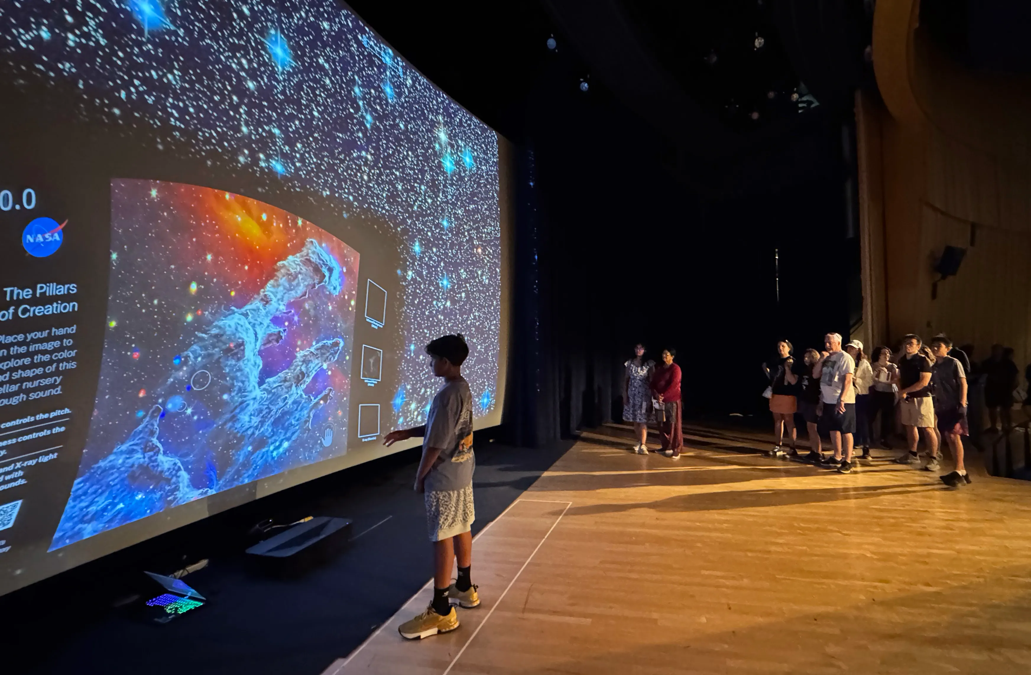 A young boy standing on stage in front of the pillars fo creation with his arm raised as a crowd looks on.